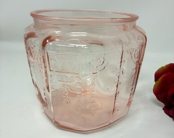 May include: A translucent pink glass jar featuring a wide circular opening and a faceted body. The exterior surface is decorated with embossed geometric patterns and scrollwork designs, creating a textured appearance across the glass panels.
