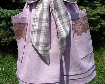 May include: A lilac-colored apron with two pockets trimmed with brown lace. The apron features a large plaid bow in shades of brown, white, and gray. The apron is displayed on a wooden form.