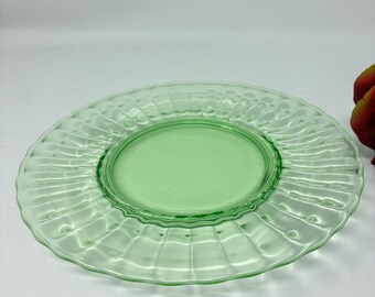 May include: A round, light green glass plate featuring a smooth, flat center surrounded by a wide, textured rim with a radial, ribbed pattern. The translucent glass has a distinct pale green hue and a scalloped edge, creating a decorative effect.
