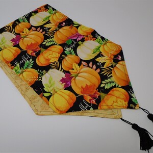 May include: A decorative table runner featuring a black background with orange and white pumpkins, fall leaves, and the words "thankful" and "blessed". The runner has a tan border and black tassels.