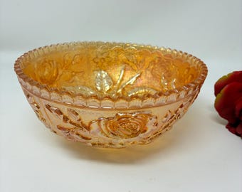 May include: An iridescent marigold orange glass bowl featuring a raised relief pattern of blooming roses and leaves on the exterior. The bowl has a scalloped, serrated rim and a reflective, metallic finish that catches the light across its surface.