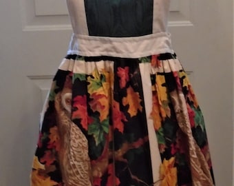 May include: An off-white apron with a dark green rectangular panel on the bib. The skirt features a colorful autumn leaf and owl print on a black background. The apron has a white ruffle trim.