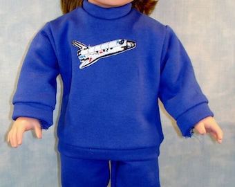 May include: A doll wearing a blue sweatshirt and matching pants, with a white and black embroidered space shuttle design on the chest. The doll is also wearing white sneakers. The doll has brown hair and is standing in front of a light blue background.
