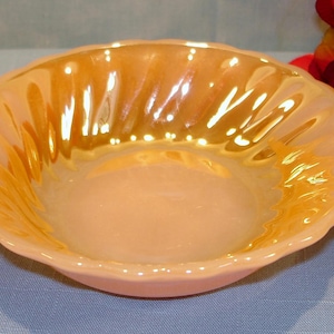 May include: A round, peach-colored glass bowl with an iridescent finish. The bowl features a scalloped edge and a series of vertical, fluted ridges along the interior walls that create a textured, light-reflecting surface throughout the piece.