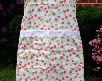 May include: A white apron with a floral pattern of red flowers and green leaves. The apron has a white lace trim on the pockets and a matching floral bow at the neck. It is displayed on a mannequin.