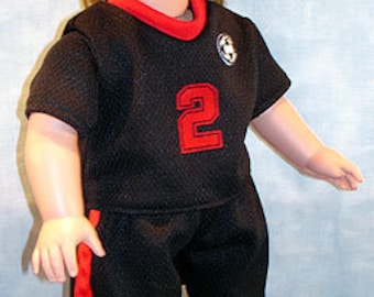 Puede incluir: Muñeca con uniforme de fútbol negro con detalles rojos, incluyendo una camiseta con el número 2. La muñeca está de pie sobre un balón de fútbol, con calcetines y pantalones cortos rojos. La muñeca tiene cabello castaño.