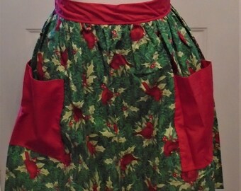May include: A festive apron with a red waistband and pockets. The apron's main fabric is green with a pattern of red cardinals and holly leaves. The apron is designed to be worn around the waist.