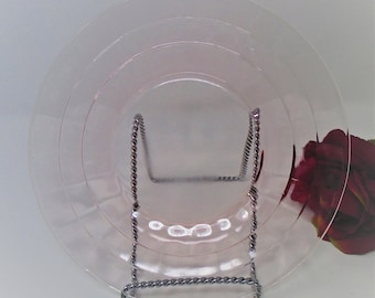 May include: A translucent pale pink glass plate featuring a series of concentric circular ridges on its surface. The plate is held upright by a silver-toned metal display stand with a twisted rope-like texture along its frame.