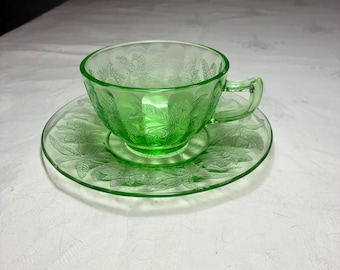 May include: A translucent light green glass teacup and matching saucer set. The cup and saucer feature a molded relief pattern of leaves and floral elements. The glass has a vibrant, bright green hue and a smooth, glossy finish throughout.