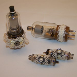 May include: Three vintage glass vacuum tubes with decorative lace and pearl accents. The tubes are different sizes and have a brown, clear, and silver finish.