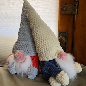 May include: Two crocheted gnomes, one with a gray and orange hat and the other with a white and blue hat. Both gnomes have white beards and pink noses.