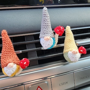 May include: Three crocheted gnome ornaments with red mushroom accents. The gnomes are hanging from a car's dashboard.