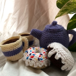 May include: A crocheted tea set with a purple teapot, two cream-colored cups, and two donuts. One donut is white with colorful sprinkles and the other is brown with white frosting.