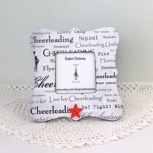 May include: White picture frame with a black and white design featuring the word "Cheerleading" repeated multiple times. The frame has a red star in the lower right corner. The frame is sitting on a white doily.