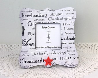 May include: White picture frame with a black and white design featuring the word "Cheerleading" repeated multiple times. The frame has a red star in the lower right corner. The frame is sitting on a white doily.