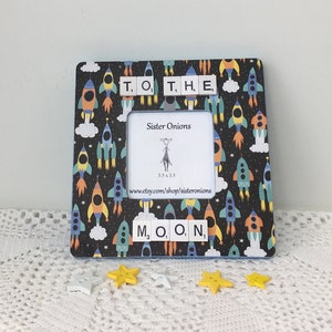 May include: A black picture frame with a rocket ship pattern. The frame has a white mat and a clear glass front. The frame is decorated with letter tiles that spell "TO THE MOON".
