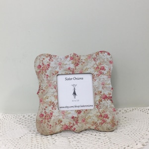 May include: A decorative photo frame with a floral pattern in pink and cream. The frame has a unique, ornate shape and includes the text "Sister Onions" and "3.5x3.5". The frame is displayed on a white, lace doily.