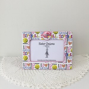 May include: A rectangular picture frame featuring a softball-themed pattern with yellow balls, hearts, and text including "CATCH," "SOFTBALL," "PITCH," "RUN," and "SLIDE." A circular button with a heart design sits on top. The center displays "Sister Onions."