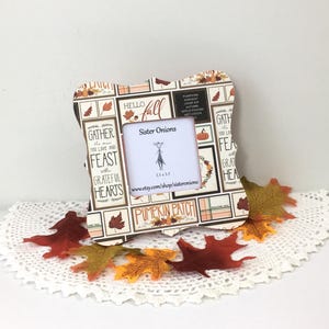May include: A square picture frame with a scalloped edge featuring a collage of autumn-themed text and illustrations including pumpkins and leaves. The center insert displays the text Sister Onions and 3.5 x 3.5 with a small line drawing.