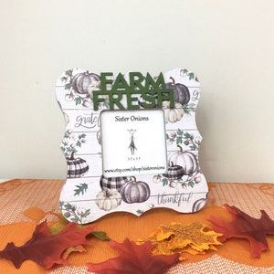 May include: A decorative photo frame with a fall theme. The frame features a white wooden background with pumpkin and cotton illustrations. The green text on the frame reads "FARM FRESH". The frame is 3.5 x 3.5 inches.