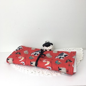 May include: A rolled-up red fabric item with a black ribbon, featuring a pattern of sheep, chickens, and other farm animals. A small sheep figurine sits on top. The item is on a white lace doily.