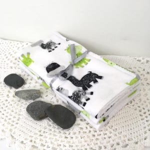 May include: A stack of white fabric cloths featuring a repeating pattern of black and lime green stylized llama illustrations. The cloths are folded into a rectangular shape and tied together with a thin, light gray satin ribbon.