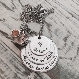 May include: Silver necklace with three layered discs. The top disc says "Briana" with a heart above the name. The middle disc says "Class of 2016". The bottom disc says "Master Social Work". A small silver charm with a graduation cap and a brown gemstone hangs from the top of the necklace.