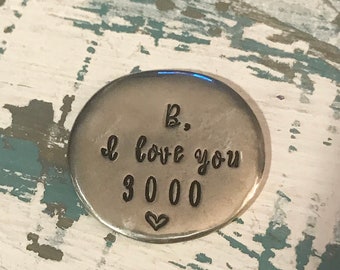 May include: A small, silver-colored, round metal token with the phrase "B, I love you 3000" and a heart symbol. The token is set against a distressed, white and blue painted wooden surface.
