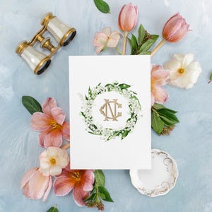 Könnte beinhalten: Eine weiße Karte mit einem goldenen Monogramm "NE", umgeben von einem Kranz aus weißen Blüten und grünen Blättern. Die Karte ist umgeben von rosa Blüten, grünen Blättern und einem Paar antiker Operngläser.