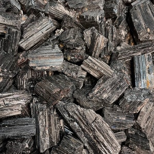 May include: A close-up view of a pile of raw, black tourmaline crystals. The crystals are rough-hewn, with a dark, metallic sheen. The crystals are in various sizes and shapes, creating a textured, natural appearance. The overall aesthetic is earthy and mineral-focused.