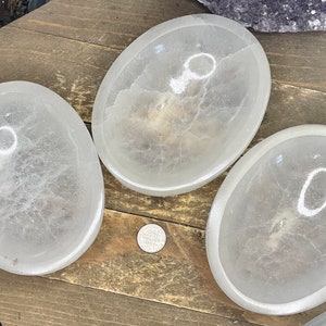 May include: Three oval-shaped, translucent white selenite bowls are arranged on a wooden surface. The bowls have a smooth, polished appearance and a shallow, concave design. A coin is placed near the bowls.