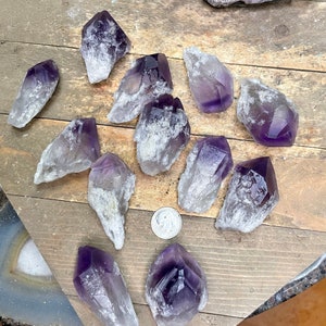 May include: A collection of amethyst crystal points with varying shades of purple and white. The crystals are arranged on a wooden surface.