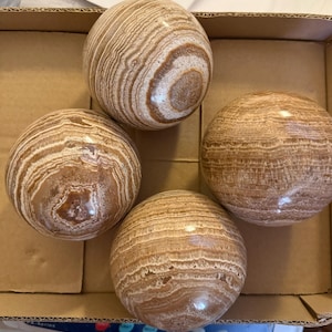 May include: Four decorative spheres with a banded, swirling pattern in shades of brown and cream. The spheres are smooth and polished, with a natural stone appearance. They are arranged inside a cardboard box.