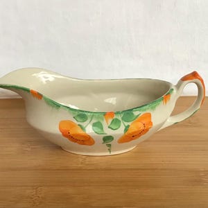 May include: A cream-coloured ceramic gravy boat featuring a hand-painted design of bright orange poppy flowers with green leaves. The vessel has a curved pouring spout, a sturdy handle, and a green painted rim that accents the top edge.