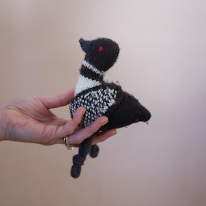 May include: A handmade knitted bird toy in black, white, and gray. The bird has a black head with red eyes, a white and black striped neck, and a gray tail and feet. The toy is held in a hand.