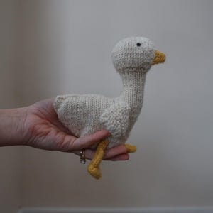 May include: A cream-coloured knitted goose toy with a yellow beak and feet. The goose has a long neck and small black eyes. The toy is held in a person's hand.