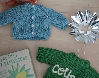 May include: A miniature blue knit cardigan with white buttons and a miniature green knit sweater with the name Colby embroidered in white yarn. A silver metallic star-shaped reflector and a box labeled Faceted Light Reflectors are also present.