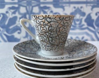 May include: A white porcelain teacup and a stack of three matching saucers, each decorated with a delicate black and gold dotted swirling pattern. The teacup features a gold-trimmed rim and a curved handle, resting atop the saucers.