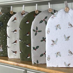 May include: A selection of tea cosies featuring festive designs such as deer, robins, holly, and owls. Each cosy has a loop and wooden button. The cosies are displayed on a wooden surface.