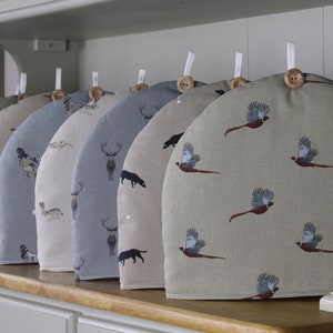 May include: A set of seven tea cosy covers featuring various animal designs. The covers are crafted from a light-coloured fabric with a subtle texture. The designs include a pheasant, a deer, a rabbit, a hen, a cat, and a dog.