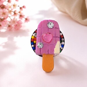Peut inclure: Une bague ornée d'une breloque en forme de glace à l'eau violette avec un motif quadrillé, un strass rose en forme de cœur, deux strass ronds transparents et une petite touche de glaçage blanc sur une base circulaire parsemée de perles colorées.