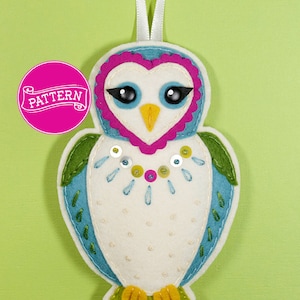 Puede incluir: Un adorno de búho de fieltro con cuerpo blanco, alas azules, patas verdes y una cara en forma de corazón rosa con ojos negros y un pico amarillo. El búho está decorado con lentejuelas y cuentas.