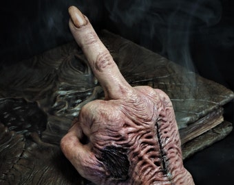 May include: A detailed, realistic hand sculpture with a fleshy, textured surface. The hand is giving the middle finger, with the index finger extended upwards. The hand is resting on a dark brown, textured book.