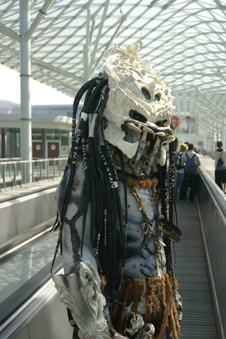 Pu&ograve; includere: Una maschera bianca da Predator con dreadlock e un materiale marrone simile alla pelliccia intorno al collo. La maschera ha un design dettagliato con una mascella aperta che mostra i denti.