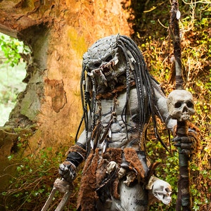 Pu&ograve; includere: Un costume da Predator con una maschera grigia e nera, dreadlocks e un perizoma di pelliccia. La figura tiene una lancia sormontata da un teschio e un paio di lame nell'altra mano.