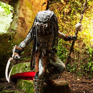 Pu&ograve; includere: Un costume da Predator con una maschera dettagliata, dreadlocks e una lancia sormontata da un teschio. Il costume &egrave; indossato da una persona in piedi in un ambiente boschivo.