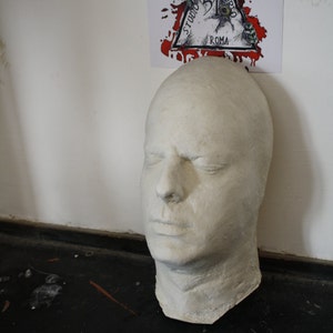 Può includere: Una maschera di gesso bianco di un viso umano. La maschera è appoggiata su una superficie nera. Gli occhi sono chiusi e la bocca è leggermente aperta.