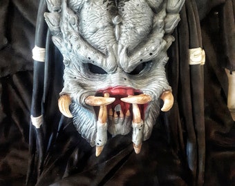 May include: A gray and black Predator mask with red detailing around the mouth and sharp teeth. The mask is made of plastic and has a textured surface.