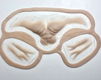 Puede incluir: Un molde de silicona beige con tres secciones en relieve, una grande y dos más pequeñas, que se asemejan a la piel humana con arrugas. El molde está etiquetado como "CHESTAF-85A" y "LEFT".