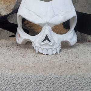 May include: A white skull mask with a cracked surface and a black strap. The mask has eye sockets, a nose cavity, and a row of teeth. A small red mark is visible on the left side of the mask.